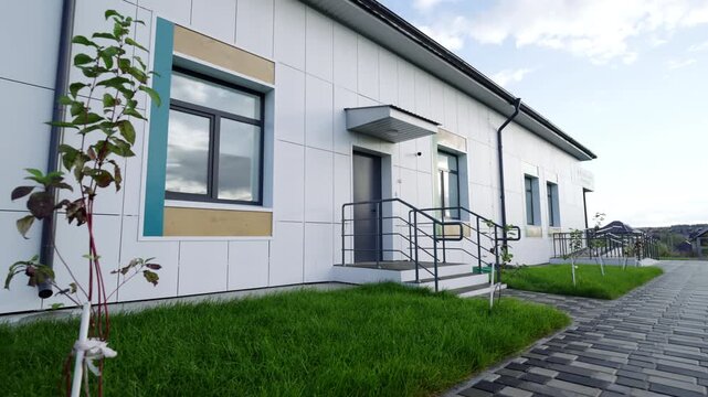Modern building shows white cladding dark windows and paved walkway. Fresh green grass under cloudy sky creates professional atmosphere