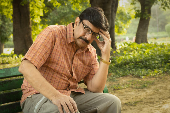 Thoughtful Indian mid aged man sitting alone on a park bench with a serious and worried expression. Concept of stress, anxiety, loneliness, mental health, retirement challenges, financial concerns.