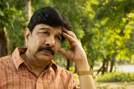 Indian senior man experiencing a sudden severe headache while sitting on a park bench. Outdoor healthcare concept highlighting head pain, migraine symptoms, stress, senior health, neurological problem