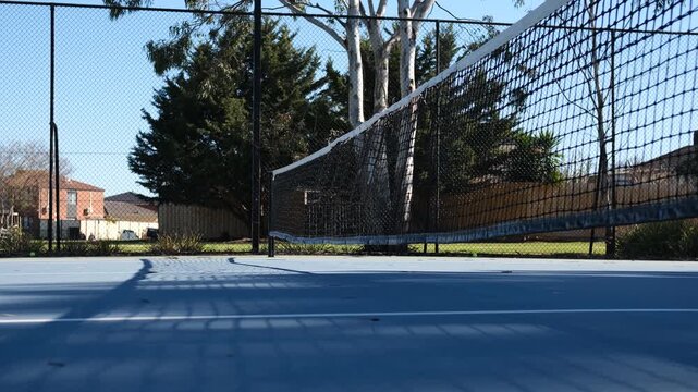 A blue hardcourt suburban tennis court near some Australian homes. The surface reflects shadows cast by the net and surrounding tall eucalyptus trees, a chain-link enclosure.