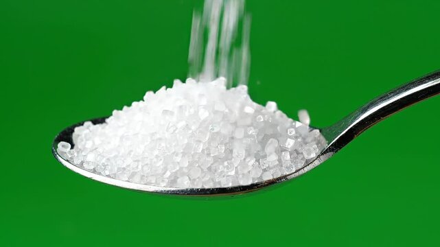 Close-up of white salt crystals pouring from a metal spoon on green background