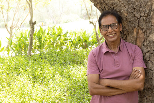 Happy Indian senior citizen smiling confidently with crossed arms in a lush green park. Portrait of a cheerful elderly man wearing eyeglasses, enjoying an active and healthy retirement lifestyle in a 