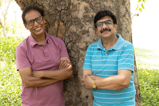 Two happy Indian senior citizens standing together with folded arms in a public park, smiling at the camera. Portrait of elderly male friends enjoying companionship, active retirement, friendship.