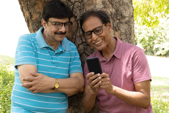 Happy Indian senior man looking at a smartphone while spending time outdoors in a green park. Concept of digital lifestyle, mobile technology, online communication, social media, internet access.