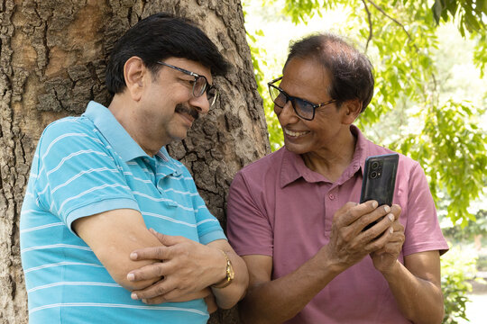Happy Indian senior man looking at a smartphone while spending time outdoors in a green park. Concept of digital lifestyle, mobile technology, online communication, social media, internet access.