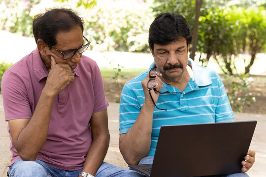 Two Indian senior citizens using a laptop together while sitting in a public park. Elderly men learning digital technology, browsing online, and discussing information, highlighting digital literacy, 