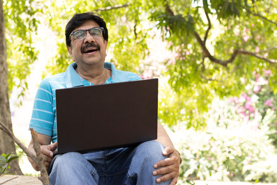 Happy Indian senior man using a laptop while sitting outdoors in a lush green park, smiling and enjoying digital connectivity. Concept of active aging, remote work, online learning, technology.