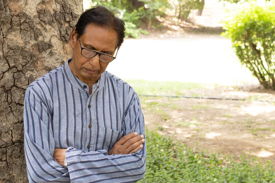 Sad Indian senior citizen sitting alone in a public park, looking thoughtful and emotional. Mature man wearing eyeglasses and traditional casual clothing, expressing loneliness, depression, aging.