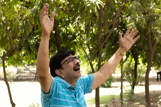 Happy Indian senior citizen standing with open arms in a lush green public park, enjoying nature and an active retirement lifestyle. Smiling elderly man wearing eyeglasses, expressing freedom.