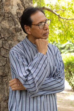 Sad Indian senior citizen sitting alone in a public park, looking thoughtful and emotional. Mature man wearing eyeglasses and traditional casual clothing, expressing loneliness, depression, aging.