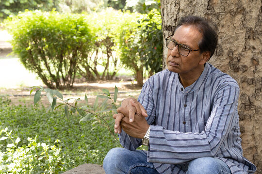 Sad Indian senior citizen sitting alone in a public park, looking thoughtful and emotional. Mature man wearing eyeglasses and traditional casual clothing, expressing loneliness, depression, aging.