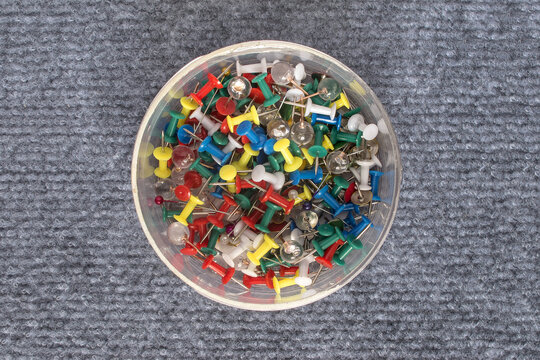A set of push pins in a round clear plastic box. Plastic heads in various colors (red, yellow, green, blue, white) and transparent options, with a metal pin. Isolate on a gray textured background.