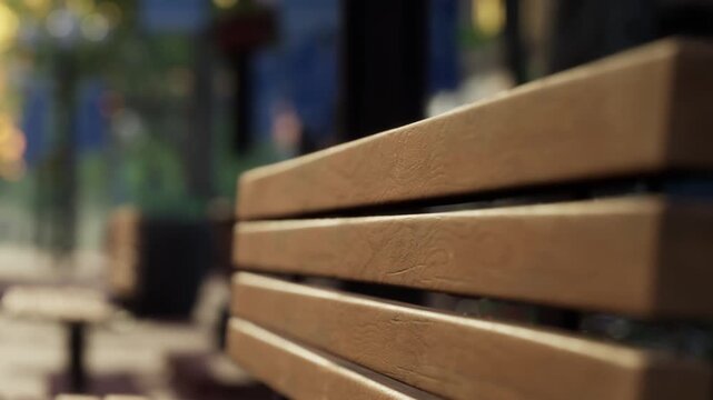A wooden bench sits in a city park. The bench has slats and shows sunlight shining down. People walk in the background, enjoying a sunny day in the park.