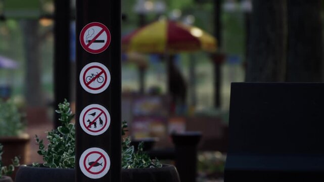 A sign stands in a park showing rules for visitors. The sign has symbols indicating no smoking, no bicycles, no pets, and no littering. People can be seen in the background enjoying the park.