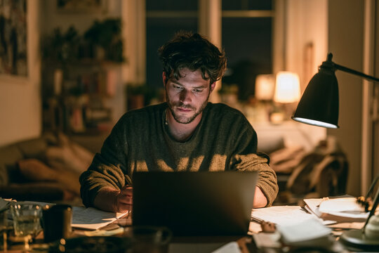 Focused Young Man Working Late at Night on a Laptop in a Cozy Dimly Lit Home Office, Representing Remote Work, Dedication, Freelance Lifestyle, Productivity, and Deep Concentration in a Quiet