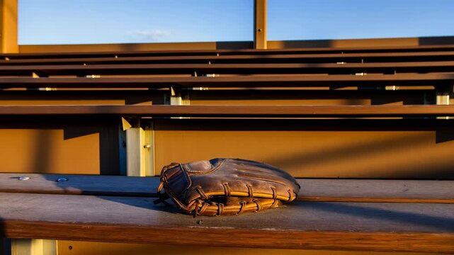 Worn leather baseball glove resting on weathered wooden bench inside empty stadium bleachers under warm sunlight, rich texture, vintage sports mood, quiet outdoor setting
