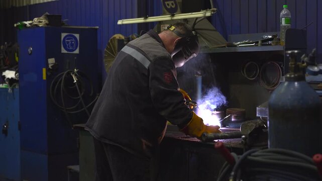 Worker welding metal parts on heavy workbench in industrial workshop with bright arc light, flying sparks, smoke, protective gear, and rugged fabrication tools, with welder, welding, worker, torch