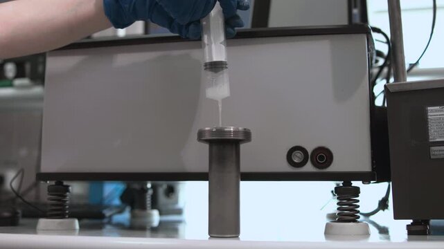 Gloved hand dispenses thin liquid into threaded metal fitting beside benchtop machine, then withdraws syringe during precise industrial assembly on work surface