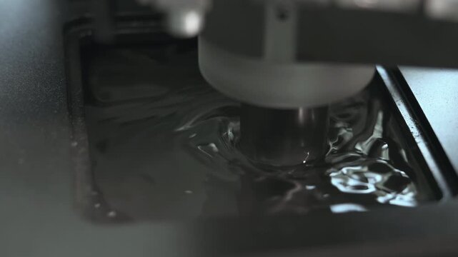 Metal probe touching dark liquid in shallow tray, creating concentric ripples, bright reflections, and subtle surface distortion under low-key industrial lighting