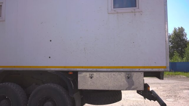 Parked white box truck rear view with dual wheels, yellow reflective stripe, mudflaps, cargo body, and heavy duty undercarriage on bright unpaved lot, with truck, cargo, box, wheel detail