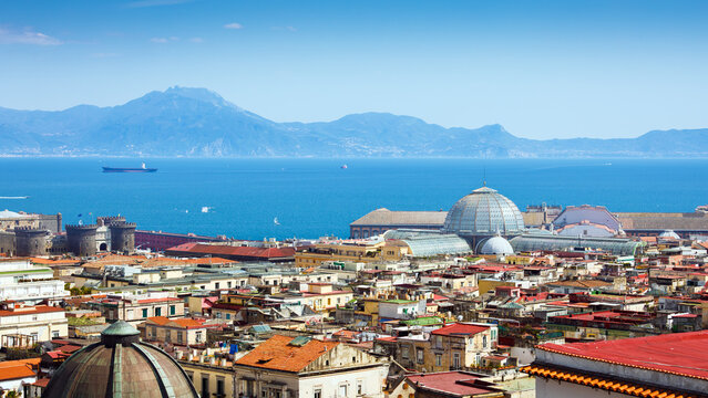Naples panorama overlooks Gulf of Naples beneath distant coastal mountains. Naples rooftops, Castel Nuovo, Galleria Umberto I, and calm blue water extend under clear summer sky across southern Italy