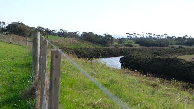 Fence lines overlooking Hovells Creek as it winds through green rural countryside near Corio, Australia, with rolling hills and busy traffic on country highway on horizon. A quiet green reserve 