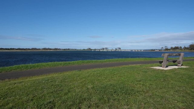 A wooden bench overlooks a wide, calm waterway at Hovells Creek, Corio Australia,  on a grassy foreshore with a walking path. A peaceful, open green coastal reserve.