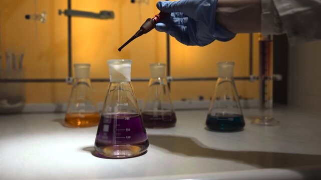 Gloved hand carefully releases purple liquid from glass dropper above Erlenmeyer flask on laboratory bench, showing precise chemistry work with glassware, reflections, and focused close-up detail
