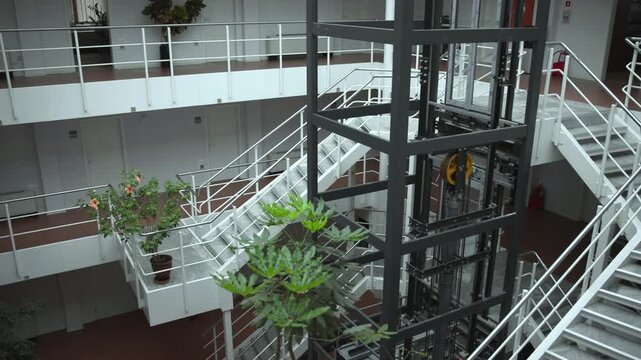 Interior atrium with exposed elevator mast, stair circulation, railings, balconies, terracotta floor, and potted greenery from elevated oblique view, with atrium, elevator, mast, framework detail