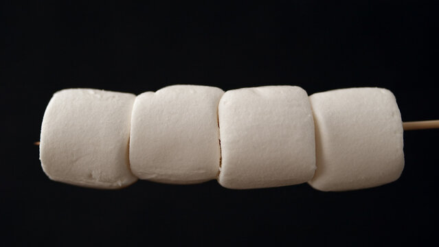 Four marshmallows threaded onto a wooden stick, arranged in a row against a solid black background.

