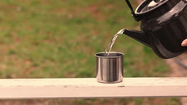 Water poured from kettle into simple metal cup on empty table representing everyday life, travel atmosphere and basic human needs with focus on hydration, modest objects and authentic outdoor environm