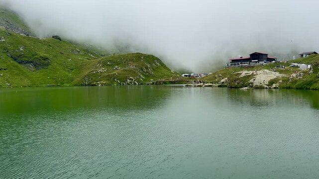 Panoramic view of Balea Lake in the Romanian Carpathian Mountains with calm alpine water, mountain chalet, rocky shoreline, green slopes and low clouds creating a peaceful natural landscape