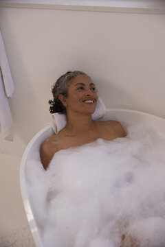Senior African American woman reclining in bathtub at home, smiling, foam and towel headrest