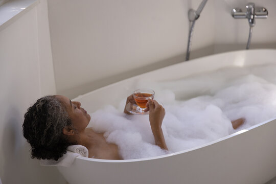 Senior African American woman resting in home bathtub with foam, holding glass teacup and saucer