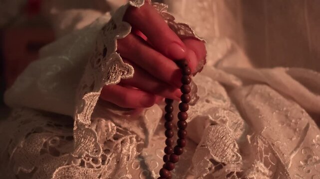 Close up slow motion video of a Muslim woman sitting in white embroidered mukena holding prayer beads gently during evening dhikr.

