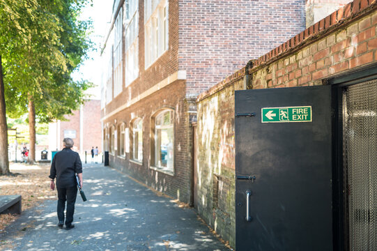 Shallow focus of an opened Fire Exit door at the rear of a restaurant kitchen in the UK. A member of the public can be seen walking adjacent to a city park in the town of Ely, UK.