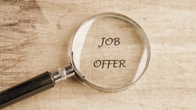 A person uses a magnifying glass to read a hidden job offer on a wooden table. The letters appear more clearly as the glass is moved closer