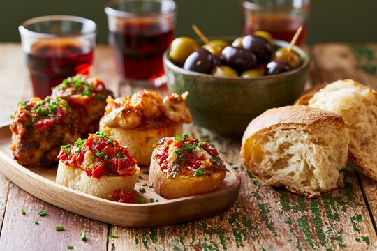 Tapas spread featuring pintxos with boqueron, olives, and bread on a rustic wooden table, accompanied by glasses of red beverage in the background