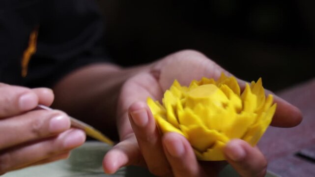Close up human hand carving yellow fruit into intricate flower shape using small carving knife in Bangkok Thailand.