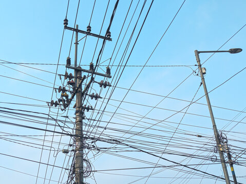 A complex network of overhead electrical power lines and telecommunication cables tangled around concrete utility poles under a clear blue sky. The image illustrates urban infrastructure, electricity 