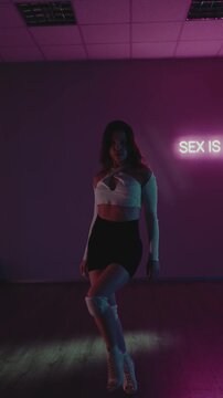 A young woman dances seductively in a dimly lit studio with pink and green lighting. She wears a white crop top, black shorts, and knee pads, moving rhythmically to the music.