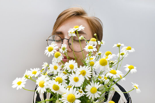 Young woman with round glasses smelling fresh chamomile flowers on gray background. Lifestyle portrait with white daisies, romantic mood, summer gift, birthday celebration, floral arrangement design.