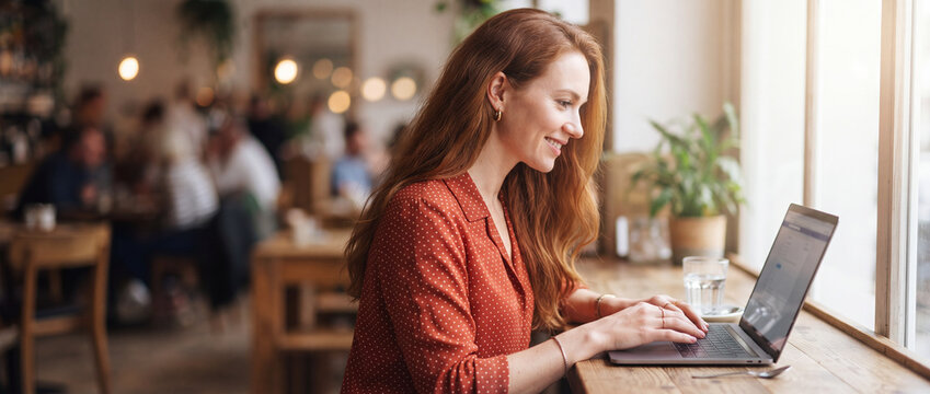 Woman working on laptop in cafe with warm natural window light, wearing a polka dot blouse while freelancing, browsing, and studying online in a cozy indoor setting for commercial technology