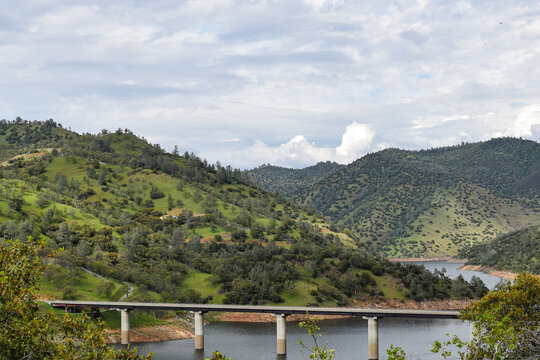 A bridge spans a reservoir surrounded by rolling green hills along California State Route 120, a scenic route toward Yosemite National Park. Dramatic clouds and lush spring vegetation create a strikin