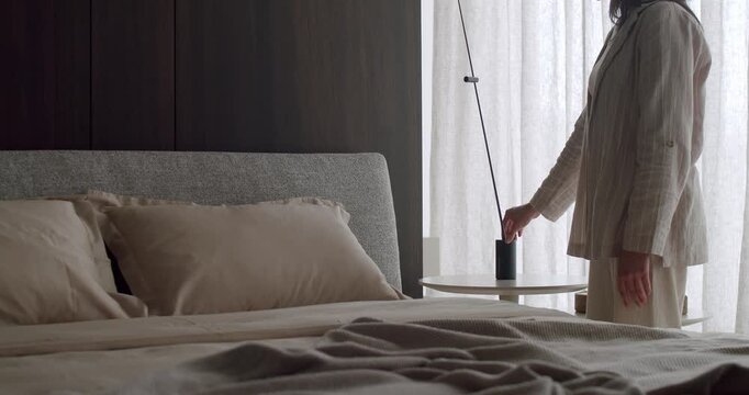 A woman in a cozy bedroom turning off a lamp, creating a peaceful and calm atmosphere with beige bedding and soft lighting. Woman Turning Off Lamp In Cozy Bedroom With Beige Bedding