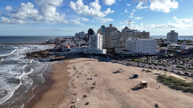 Drone footage flying along Playa Mansa with sandy beaches, gentle waves, the Punta del Este skyline and coastal architecture under a bright blue sky in Uruguay