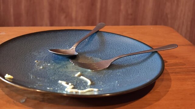 Slow motion wide shot of an empty dirty ceramic plate with crossed spoon and fork after finishing a delicious meal.

