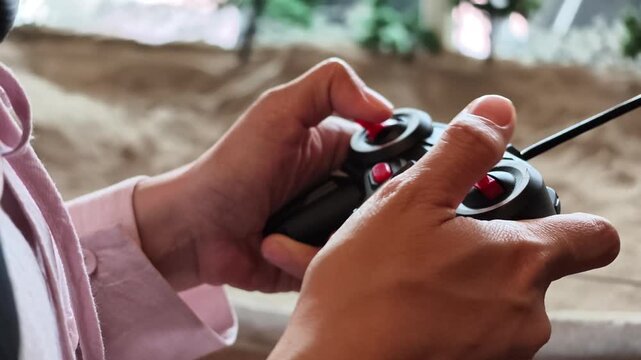 Slow motion angle view of hands holding a black radio-control transmitter with red joysticks in front of an off-road track.
