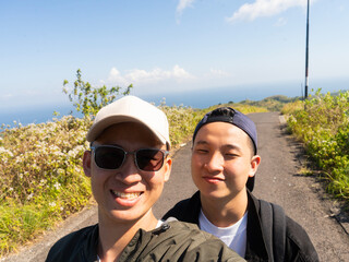 Two friends smile on a scenic coastal path. Wearing hats and sunglasses, they enjoy the view. Bright sky and distant sea frame the moment. Perfect for travel, adventure, or friendship themes © Freddy Chandra