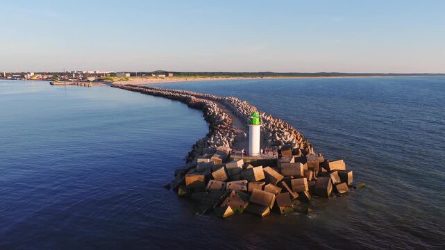Aerial view of Ventspils, Latvia, shows curved tetrapod breakwater to a white lighthouse with green lantern, sandy beach, woods, port tanks, and people on the pier.
